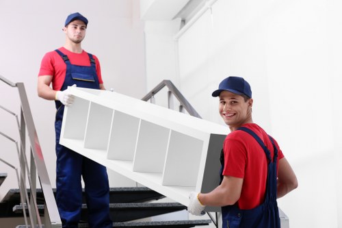 Two professional movers from Chiswick Man with Van are carrying a white, open-fronted shelving unit made of wood or particleboard, with a smooth finish. The mover in the foreground, smiling and wearing a navy blue cap, red shirt, and dark blue overalls, is holding the shelving unit at the middle and right side, ensuring stability. The second mover, standing on the staircase behind him and wearing a matching outfit, is grasping the left side of the shelving unit and appears to be carefully supporting it during the move. They are positioned on a residential staircase with dark grey carpeted steps and metal handrails, inside a bright, modern house with white walls and natural lighting. The environment suggests an interior space prepared for a house relocation, aligning with professional removals services. The movers are equipped with white gloves to protect the furniture and ensure safe handling, and the scene conveys a careful, organized approach typical of house removals in the Chiswick or broader London area.