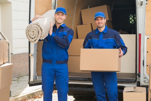 Two professional removal men dressed in blue uniforms are standing outside a residential property with an open removals van in the background. The man on the left is smiling and holding a large rolled-up carpet on his shoulder, while the man on the right is also smiling and holding a medium-sized cardboard box. Behind them, the van's interior is visible, filled with stacked cardboard boxes of various sizes and some furniture wrapped in protective material. The scene takes place on a residential street near a driveway, with a glimpse of a house wall and pavement visible. The environment appears bright and clear, indicating daytime. The image illustrates house removals services in the Chiswick area, highlighting the process of loading household items into a professional removal vehicle in preparation for a move, with attention to the careful handling of textiles, boxes, and furniture as part of a typical house relocation service by Chiswick Man with Van.