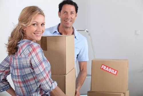 A smiling man and woman in a bright, modern interior are engaged in moving house, with the woman holding a cardboard box and the man standing behind her next to a stack of similarly packed boxes. The woman has long, wavy blond hair and is dressed in a casual, plaid shirt, while the man has short dark hair and is wearing a light blue shirt. The boxes are made of brown cardboard, some with visible labels, one marked as 'FRAGILE,' indicating they are packed for a removal. In the background, additional boxes are visible, along with a plain white wall and a partially visible metal staircase or railing. The setting suggests an organized removal process in a residential space, likely in or near Chiswick, as indicated by the website's context, with the focus on careful packing and handling of household items in preparation for a move. The lighting is natural, giving the scene a warm, inviting atmosphere consistent with house removals in London suburbs.