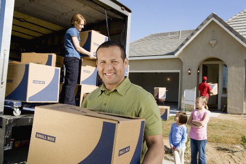 A group of household movers is actively engaged in the process of relocating belongings from a residential property in Chiswick. In the foreground, a man with short dark hair, wearing a green shirt, is smiling while holding a medium-sized cardboard box labeled 'Small Box.' Behind him, a woman with her hair tied back, dressed in a blue top, is standing inside an open removals truck, lifting or placing another cardboard box on the vehicle's floor. To the right of the image, three young children, two girls and a boy, are standing on the lawn outside a modern, light-colored house with a tiled roof and a small porch; they appear to be observing the activities. In the background, a person dressed in red is near the pathway leading to the house, and the driveway is visible with a neatly maintained lawn and garden. The scene is set on a sunny day, providing natural light that highlights the packed boxes, the textures of the cardboard, and the exterior details of the home, reflecting the professional house removals services of Chiswick Man with Van.